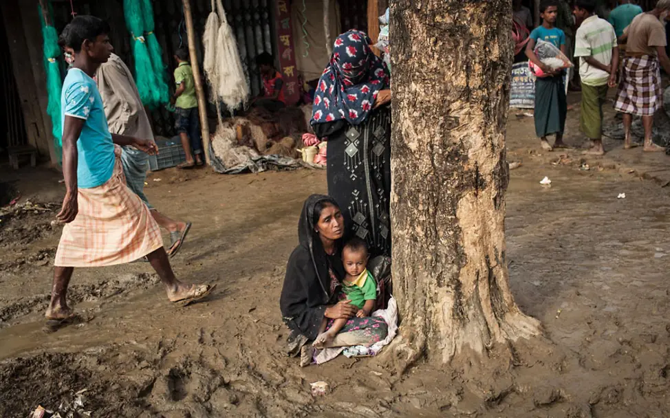 Eine Mutter hält ihr Kind in der Nähe einer Flüchtlignsunterkunft | © UNICEF/Brown Eine Mutter hat ihr Kind auf dem Schoß während sie im Schlamm sitzt