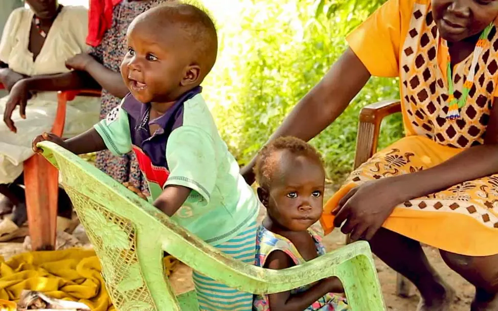 Akot hat wieder richtig viel Energie. Seine Mutter passt gut auf ihn auf. | © UNICEF/Wilson Das Baby Akot steht breit lächelnd auf einem Stuhl und hält sich an der Lehne fest.