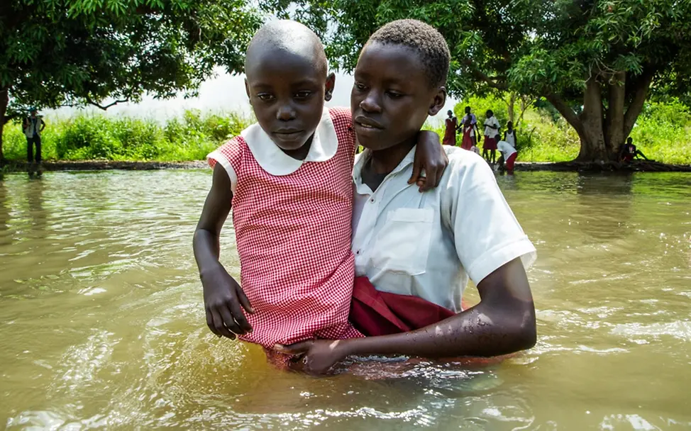 Südsudan 2021: Auf dem Nachhauseweg von der Schule hilft eine ältere Schülerin einer jüngeren. | © UNICEF/UN0540659/Chol Südsudan: Eine ältere Schülerin trägt ein jüngeres Mädchen durch das Hochwasser