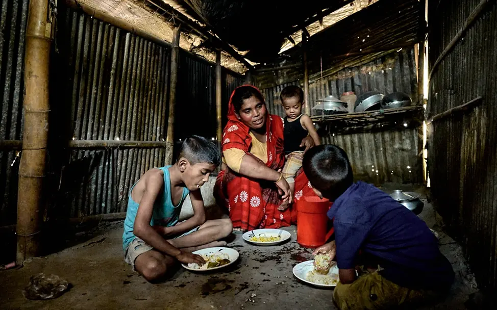 Bangladesch 2023: Atika und ihre drei Kinder haben ihr Zuhause durch Sturm und Flut verloren. | © UNICEF/UN0824111/Map Hochwasser folgen: Mutter isst mit ihren drei Kindern auf dem Boden einer Hütte