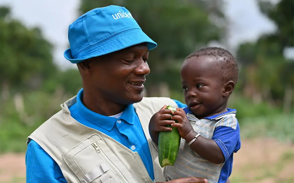 Ein UNICEF-Helfer hält ein Kleinkind auf dem Arm | © UNICEF/Dejongh Hungersnot verhindern: Ein Kleinkind mit Zucchini in der Hand bei einem UNICEF-Helfer auf dem Arm