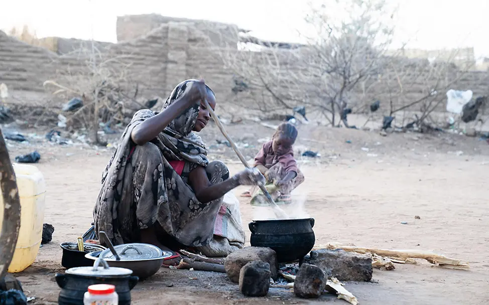 Frau kocht im Sudan am offenen Feuer. | © UNICEF/Jamal Eine Frau kocht im Sudan im Freien an einer Feuerstelle, während ein Kind in der Nähe im Staub sitzt.