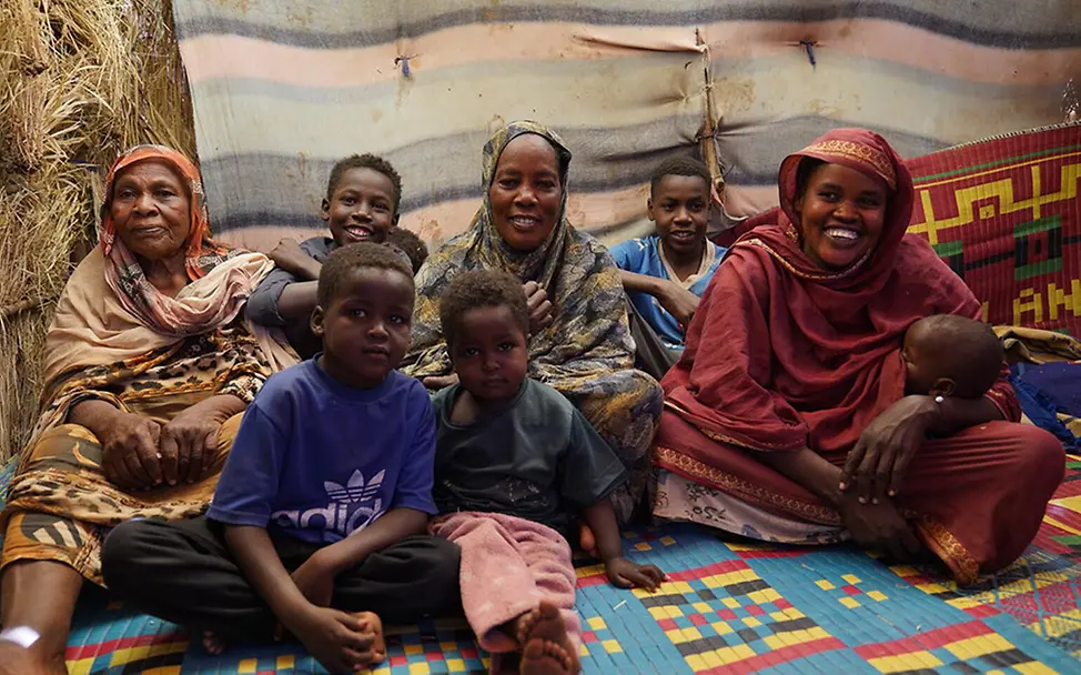 Sudan: Kinder und Frauen in einem Notunterkunftsraum | © UNICEF/Jamal Sudan: Mehrere Kinder und drei Frauen sitzen gemeinsam in einer provisorischen Unterkunft auf Matten.