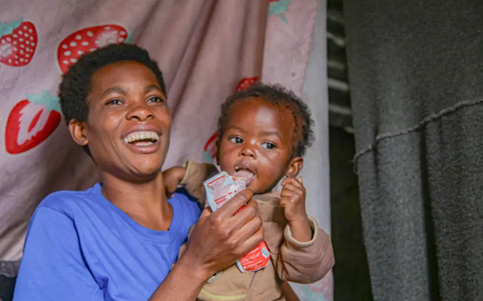 Mutter füttert ihr Kind mit lebensrettender Erdnusspaste © UNICEF/Ushindi Ein Kleinkind sitzt auf dem Arm ihrer Mutter, die es mit Erdnusspaste füttert.